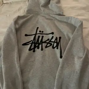 Säljer en grå hoodie från Stüssy med deras ikoniska logga i svart på både framsidan och baksidan. Tröjan har en klassisk känguruficka och justerbar huva med dragsko. Perfekt för en avslappnad stil.