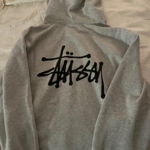 Grå hoodie från Stüssy - Säljer en grå hoodie från Stüssy med deras ikoniska logga i svart på både framsidan och baksidan. Tröjan har en klassisk känguruficka och justerbar huva med dragsko. Perfekt för en avslappnad stil.