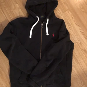 Svart hoodie från Ralph Lauren - Säljer en stilren svart hoodie från Ralph Lauren med dragkedja och vit snörning i huvan. Den har en liten röd logga på bröstet och är perfekt för en avslappnad look. Passar både till jeans och träningsbyxor.