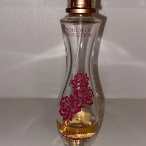 Touch of Seduction parfym - Säljer en elegant parfymflaska från Christina Aguilera, 'Touch of Seduction'. Flaskan är smal och kurvig med vackra röda blommor tryckta på glaset. Den har en gyllene kork och rymmer en doft som är både förförisk och feminin. Perfekt för speciella tillfällen eller när du vill känna dig extra glamorös.