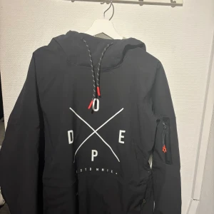 Dope Jacka - Säljer en svart jacka från Dope med tryck på framsidan. Den har en justerbar huva med dragsko och en ficka på ärmen med dragkedja. 