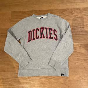 Säljer en grå sweatshirt från Dickies med röd text på bröstet. Tröjan har långa ärmar och ribbade muddar vid ärmslut och nederkant. Perfekt för en avslappnad stil.