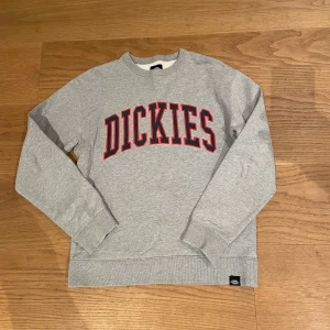 Grå sweatshirt från Dickies - Säljer en grå sweatshirt från Dickies med röd text på bröstet. Tröjan har långa ärmar och ribbade muddar vid ärmslut och nederkant. Perfekt för en avslappnad stil.