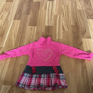 Rosa stickad klänning med hjärtmönster - Söt rosa stickad klänning med ett stort hjärta på bröstet. Den har långa ärmar och en rutig kjoldel i rosa, svart och vitt. Dekorativa knappar i midjan ger en extra touch. Perfekt för små fashionistas!