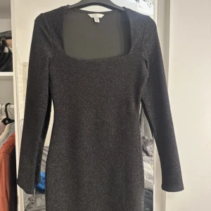 Svart glittrig klänning från KappAhl - Snygg svart glittrig klänning från KappAhl med fyrkantig urringning och långa ärmar. Perfekt för en kväll ute eller en festlig tillställning. Klänningen har en elegant och stilren design som verkligen fångar ögat.