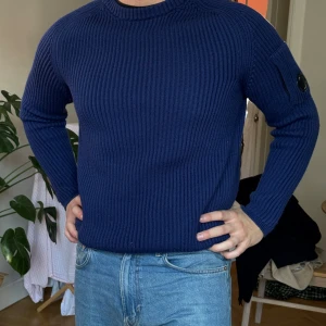 Mörkblå stickad tröja från C.P. Company - Säljer en snygg mörkblå stickad tröja från C.P. Company. Tröjan har en ribbad design och en rund halsringning. Perfekt för kyliga dagar och ger en stilren look. Märkesdetalj på ärmen för extra stilpoäng.