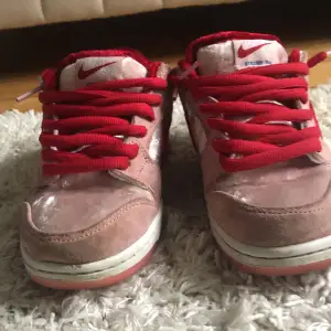 Säljer ett par Nike SB Dunk Low Strangelove i rosa och rött. Skorna har en unik design med röd sammet och rosa mocka, samt röda snören. Perfekta för dig som vill sticka ut med stil! De är i bra skick och redo för nya äventyr.