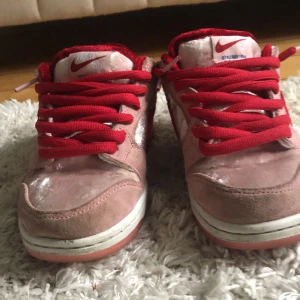 Nike SB Dunk Low Strangelove - Säljer ett par Nike SB Dunk Low Strangelove i rosa och rött. Skorna har en unik design med röd sammet och rosa mocka, samt röda snören. Perfekta för dig som vill sticka ut med stil! De är i bra skick och redo för nya äventyr.