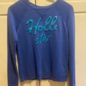 Blå tröja från Hollister - Säljer en snygg blå tröja från Hollister i storlek S. Den har långa ärmar och ett coolt glittrigt Hollister-tryck på framsidan. Perfekt för en avslappnad stil eller en chill dag ute. Tröjan är i bra skick och redo för nya äventyr! 💙