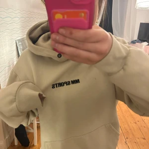 Beige hoodie från MM Sports - Säljer en beige hoodie från MM Sports med tryck på bröstet. Den har en stor och mysig huva samt långa ärmar. Perfekt för en avslappnad stil.