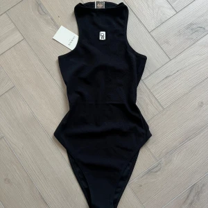 Svart baddräkt från Lis Bonne  - Ny! Snygg svart baddräkt från Lis Bonne med hög hals och ärmlös design. Perfekt för en stilren look vid poolen eller stranden. Den har en liten logotypdetalj framtill. Funkar även som body 