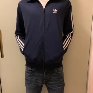 Mörkblå träningsjacka från Adidas - Säljer en snygg mörkblå träningsjacka från Adidas med de klassiska tre vita ränderna längs ärmarna. Jackan har dragkedja framtill och är i mycket bra skick. .