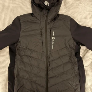 Svart Spray Hybrid Jacket från Sail Racing - Säljer en svart Spray Hybrid Jacket från Sail Racing i mycket bra skick. Jackan har en stilren design med dragkedja och långärmade ärmar. Perfekt för höst och vår med sitt quiltade mönster och syntetmaterial som håller dig varm. Den är både funktionell och snygg för vardagsbruk.