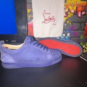 Blå sneakers från Christian Louboutin - Snygga blå sneakers i mocka från Christian Louboutin med ikoniska röda sulor. Skorna har snörning och är dekorerade med nitar på tån för en edgy look. Perfekta för att ge din outfit en lyxig touch. Storlek 39 äkta pris 4200 kr eller bud påsen fås med på bilden 