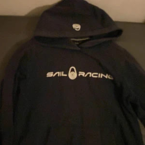Svart hoodie från Sail Racing - Snygg mörk blå hoodie från Sail Racing med logga på bröstet och huvan. Perfekt för en avslappnad stil. Den har en klassisk passform och en praktisk ficka framtill.