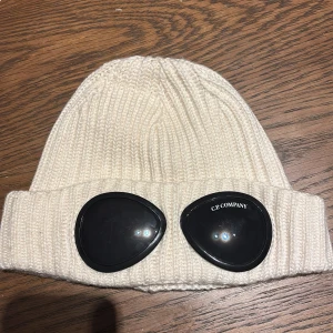 Vit stickad mössa från C.P. Company - Säljer en snygg vit stickad mössa från C.P. Company. Den har de ikoniska glasögondetaljerna framtill som ger en unik look. Perfekt nu inför vintern köpt på CP company hemsida