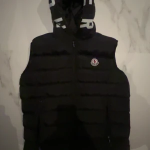 Svart dunväst med huva - Snygg svart dunväst från Moncler med dragkedja och huva. Perfekt för kyliga dagar. Märkeslogga på bröstet ger en exklusiv touch. Scan finns också 