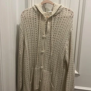 Beige stickad kofta från MANGO - Säljer en mysig cremevit stickad kofta från MANGO med huva och knappar framtill. Koftan har ett fint hålmönster och långa ärmar, perfekt för kyliga dagar. Passar bra till lager-på-lager-stil!Ny pris 599kr, använd 1 gång.  