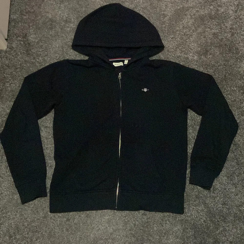 Säljer en klassisk svart hoodie från GANT i mycket bra skick. Den har en dragkedja framtill och en liten logga på bröstet. Perfekt för höst och vinter, och passar både till vardags och lite mer avslappnade tillfällen. Superbekväm och stilren! (Accepterar endast Swish pga lurendrejerier) (Checka även in andra kläder på min profil och köp tillsammans för ett rabatterat pris). Neuletakit & Villapaidat.
