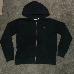 Svart hoodie från GANT - Säljer en klassisk svart hoodie från GANT i mycket bra skick. Den har en dragkedja framtill och en liten logga på bröstet. Perfekt för höst och vinter, och passar både till vardags och lite mer avslappnade tillfällen. Superbekväm och stilren! (Accepterar endast Swish pga lurendrejerier) (Checka även in andra kläder på min profil och köp tillsammans för ett rabatterat pris)