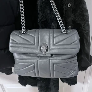 Grå kurt geiger väska - Snygg grå axelväska, den är i mycket bra skick och har en stilren design som passar till alla outfits. märket är kurt geiger och den har endast små tecken på användning! den är köpt här på plick men den är äkta