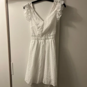 Oanvänd studentklänning från Sarapoul - Charmig vit klänning från Sarapoul med v-ringning och spetsdetaljer i midjan. Klänningen har en öppen rygg med knytning och volangärmar som ger en romantisk touch. Perfekt för sommarens festligheter!