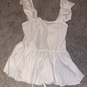 Vit topp med volangdetaljer - Söt vit topp med volangdetaljer och knytning där bak. Den har en luftig passform och är tillverkad i ett lätt material! Säljer eftersom det inte kommer till användning🤍 finns inga skador eller fläckar på den🤍
