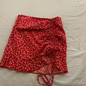Röd blommig kjol med snörning - Säljer en superfin röd kortkjol med vita blommor. Kjolen har en snygg snörning framtill som ger en cool detalj och en volangkant som gör den extra söt. Perfekt för sommardagar eller en festlig kväll!  Den är inte stretchig alls men sitter perfekt
