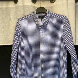 Blå och vit rutig skjorta från Ralph Lauren - Säljer en snygg blå och vit rutig skjorta från Ralph Lauren i storlek L. Skjortan är tillverkad i 100% bomull och har långa ärmar. Den har en klassisk krage och knappar framtill. Perfekt för både vardag och fest! Den är ganska liten i passformen. 💙