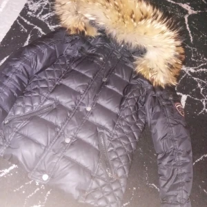 Svart dunjacka från Rockandblue - Säljer en snygg svart dunjacka från Rockandblue i storlek 34. Jackan har en fluffig pälsdetalj på huvan och quiltad design. Perfekt för kalla vinterdagar med dragkedja och knappar för extra värme. Den är i mycket bra skick och redo för nya äventyr!