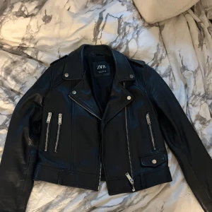 Svart skinnjacka från Zara - Säljer en snygg svart skinnjacka från Zara i mycket bra skick. Jackan har en cool bikerstil med dragkedjor och knappar. Perfekt för höst och vår, och den ger en edgy look till vilken outfit som helst. Passar både till vardags och fest!