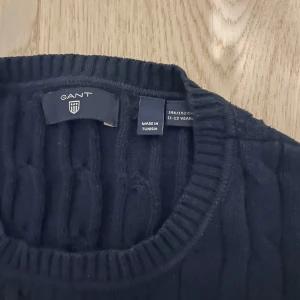 Svart kabelstickad tröja från GANT - Säljer en stilren svart kabelstickad tröja från GANT. Tröjan har en klassisk rund halsringning och långa ärmar, perfekt för kyligare dagar. Passar både till vardags och mer uppklädda tillfällen.