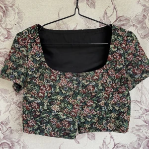 Blommig kortärmad topp - Säljer en vintageinspirerad blommig topp med korta ärmar. Toppen är croppad och har ett vackert blommönster. Köpt second hand, men inte använd av mig. (Förra säljaren på bild.) Storleken är uppskattad, se mått! Axlar (rygg): 34cm, längd: 40cm, armhåla till armhåla: 45cm, midja: 76 cm