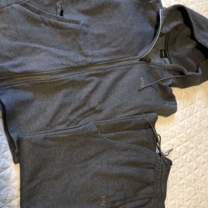 Grå sett från Boss - Säljer en stilren och bekväm grå hoodie från Boss. Den har en dragkedja framtill och en praktisk huva. Perfekt för en avslappnad stil eller träning.Även byxorna ingår i detta pris. Passar bra till både höst och vår. Hör av dig vid intresse!