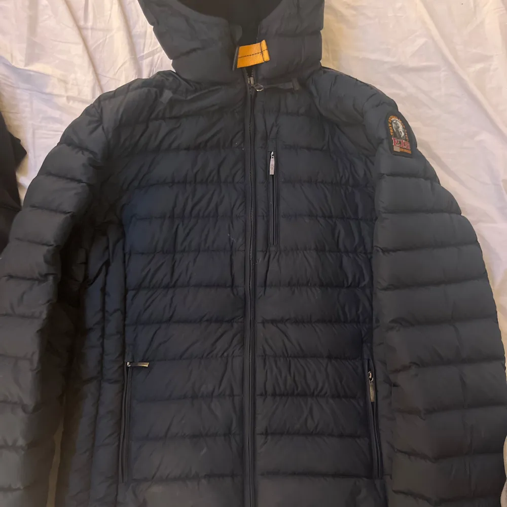 Snygg svart dunjacka från Parajumpers med huva och dragkedja. Jackan har en quiltad design och flera praktiska fickor med dragkedjor. Perfekt för kyliga dagar. Jacka har även luft hål vid armhålan. Skulle säga att jackan är i s/m Jackan har tvp små hål i vänster armen . Takit.
