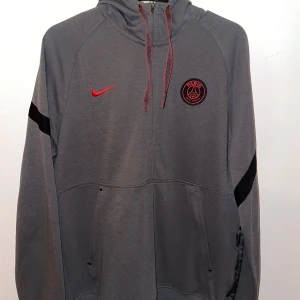 Grå hoodie från Nike med PSG-logga - Säljer en grå hoodie från Nike med PSG-logga på bröstet. Hoodien har en röd Nike-logga och snören, samt svarta detaljer på ärmarna. Perfekt för fotbollsfans! Säljer den pga den e för liten jätte bra skick! 