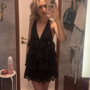 Svart glittrig klänning - Snygg svart klänning med glittriga detaljer och volanger. Den har en djup v-ringning och öppen rygg med knytband i nacken. Perfekt för en festlig kväll! Använd en gång