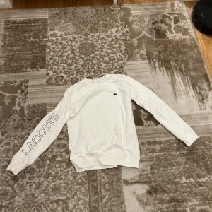 Vit sweatshirt från Lacoste - Säljer en stilren vit sweatshirt från Lacoste med deras ikoniska logga på bröstet och text på ärmen. Tröjan har långa ärmar och en klassisk rund halsringning. Perfekt för en avslappnad look.