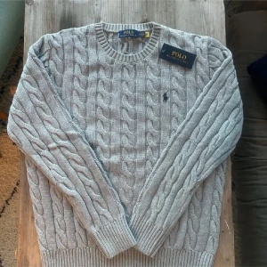   Ralph Lauren tröja (kvitto) - Säljer en helt ny stilren grå kabelstickad tröja från Polo Ralph Lauren. Tröjan har långa ärmar och en klassisk rund halsringning. Perfekt för kyligare dagar med sin mjuka och bekväma passform. 🧶