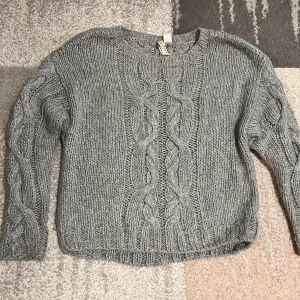 Grå stickad tröja med flätmönster - Mysig grå stickad tröja med ett snyggt flätmönster på framsidan. Tröjan har långa ärmar och en rund halsringning, perfekt för kyligare dagar. Passar bra till både jeans och kjol.