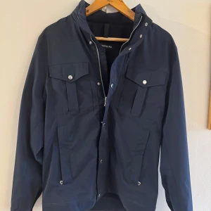 Mörkblå jacka från J.Lindeberg - Snygg mörkblå jacka från J.Lindeberg med dragkedja och knappar framtill. Jackan har flera praktiska fickor med lock och knappar. Perfekt för en stilren look.