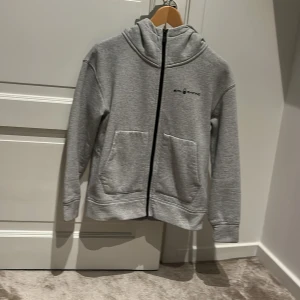 Grå hoodie från Sail Racing - Säljer en stilren grå hoodie från Sail Racing med dragkedja och två praktiska fickor framtill. Tröjan har en bekväm passform och en huva med logotypdetalj. Perfekt för en avslappnad stil. Hoodien är också väldigt skön och i mycket bra skick