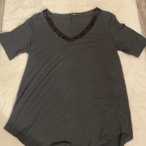 Grå t-shirt med paljettdetaljer  - Snygg grå t-shirt med v-ringning dekorerad med svarta paljetter. Perfekt för att ge en touch av glamour till din vardagslook. T-shirten har korta ärmar och en bekväm passform.