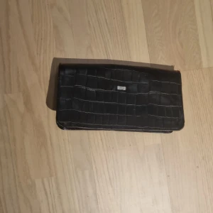 Svart kuvertväska från Kookai - Snygg svart kuvertväska från Kookai med krokodilpräglat mönster. Väskan har en elegant rosa insida i satin och stängs med en tryckknapp. Perfekt för att ge en touch av stil till din outfit.