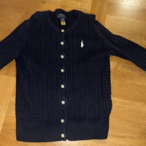 Ralph Lauren Kofta - En kofta från Ralph Lauren, köpt i USA. Säljer då den inte passet längre. Som man kan se på bilden så har den lite röd färg på armen och vid knapparna men det är knappt så att man ser. Köptes för runt 1000kr men säljer för 400kr pga. skicket. Det är en barn storlek (L/12-14)