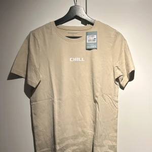 Beige t-shirt från Firefly - Säljer en beige t-shirt från Firefly i ekologisk bomull. T-shirten har texten 'CHILL' tryckt på bröstet och ett subtilt kamouflagemönster nedtill. Perfekt för en avslappnad stil.