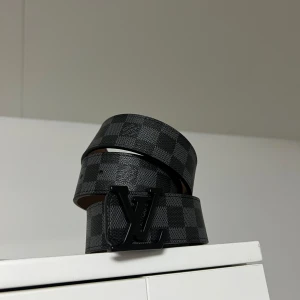 LV bälte svart - Snyggt bälte från Louis Vuitton med ett klassiskt rutigt mönster i svart och grått. Bältet har en stilren LV-logotyp som spänne och är tillverkat i skinn. Perfekt för att ge en lyxig touch till din outfit. 110cm lång vid frågor eller funderingar hör av er!