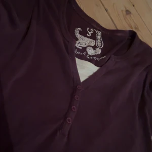 Mörklila tröja från Flash - Säljer en mörklila tröja från Flash med knappar framtill. Tröjan har en enkel design med en liten öppning vid halsen och ett tryck på insidan av kragen. Perfekt för en avslappnad stil.