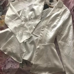 Vit satin kavaj - Elegant vit blus i satin med en figursydd passform. Den har långa ärmar och en klassisk krage. Perfekt för att ge en stilren look. Helt ny oanvänd