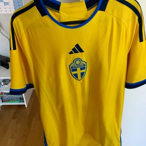 Sverige hemma ställ 22/23 - Säljer en gul svensk landslagströja från Adidas med blå detaljer och Sverigelogo på bröstet. Tröjan har korta ärmar och spelarens namn 'Isak' med nummer 9 på ryggen. Perfekt för fotbollsfans! 💙💛